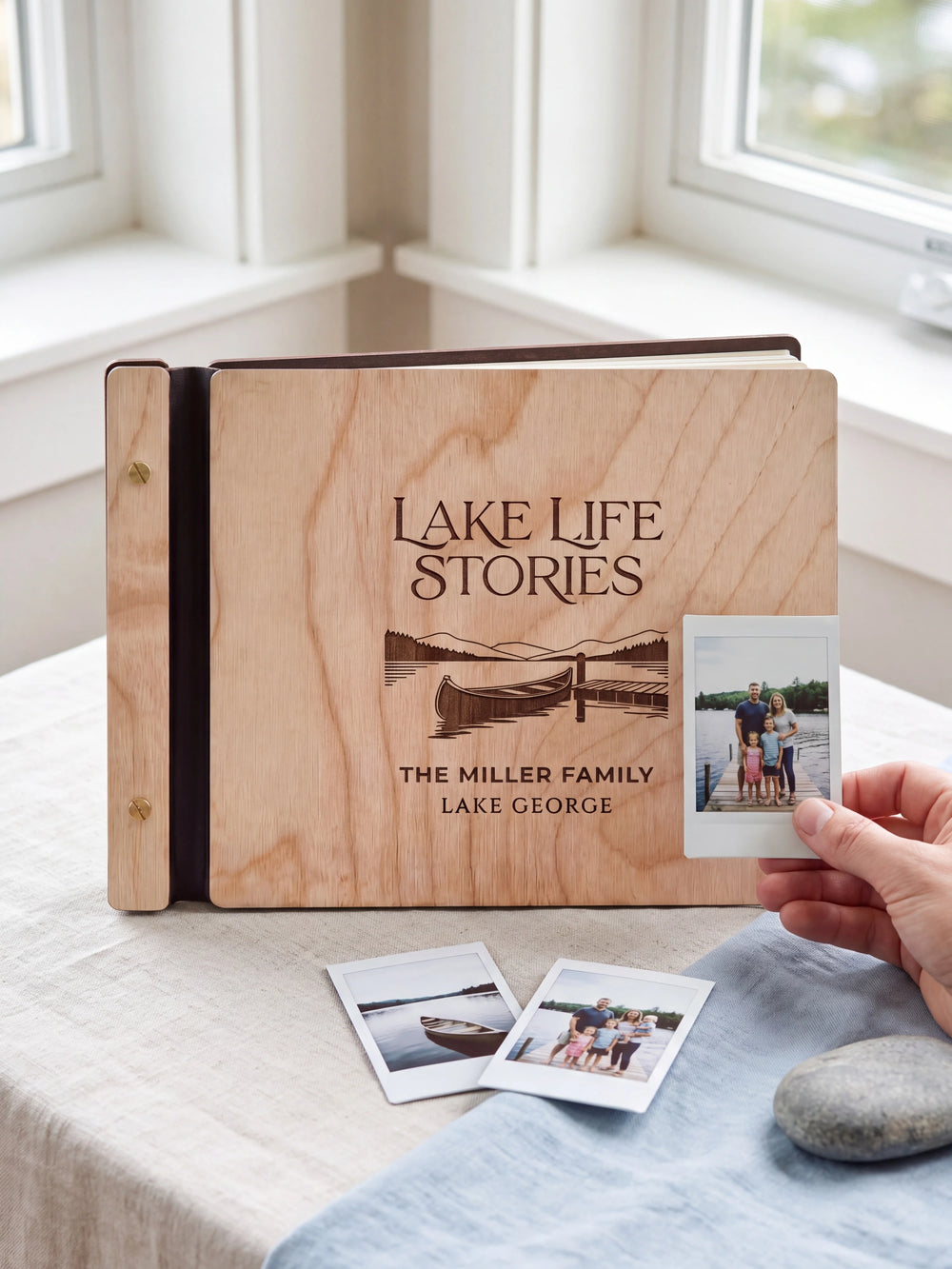 Natural wooden album with dark engraving of a canoe beside a lakeside dock. A hand holds an instant photo beside the closed cover, with loose snapshots nearby.