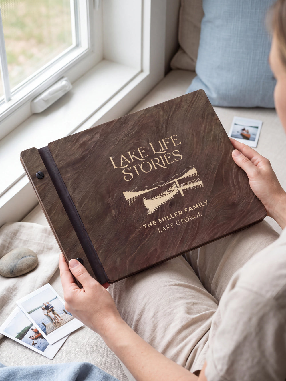 Hands hold a dark brown Jacobean wooden album close to the camera. Cream engraving shows a canoe beside a lakeside dock, personalized wording and the black binding strip.