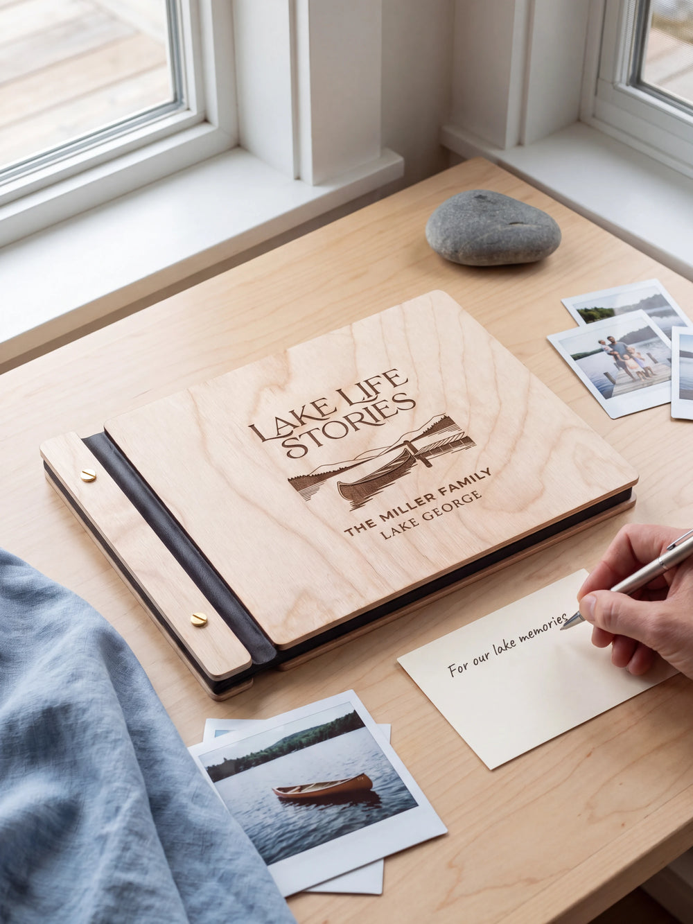 A Natural wooden album showing a canoe beside a lakeside dock rests beside a separate gift card. A hand writes 