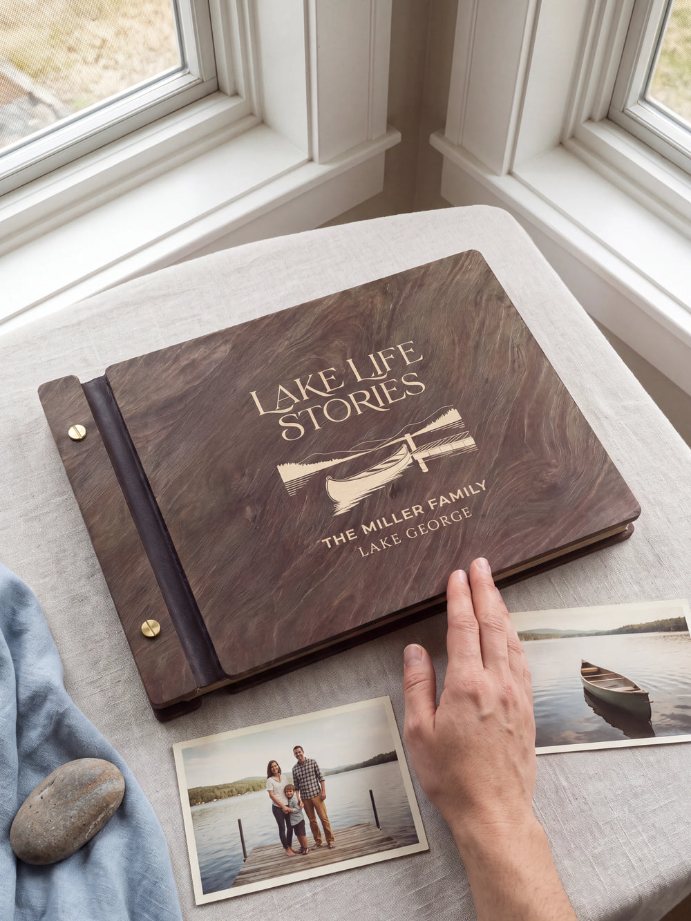 A hand rests beside the closed dark brown Jacobean album on a table. Cream engraving and personalized wording are visible, with travel photos nearby.