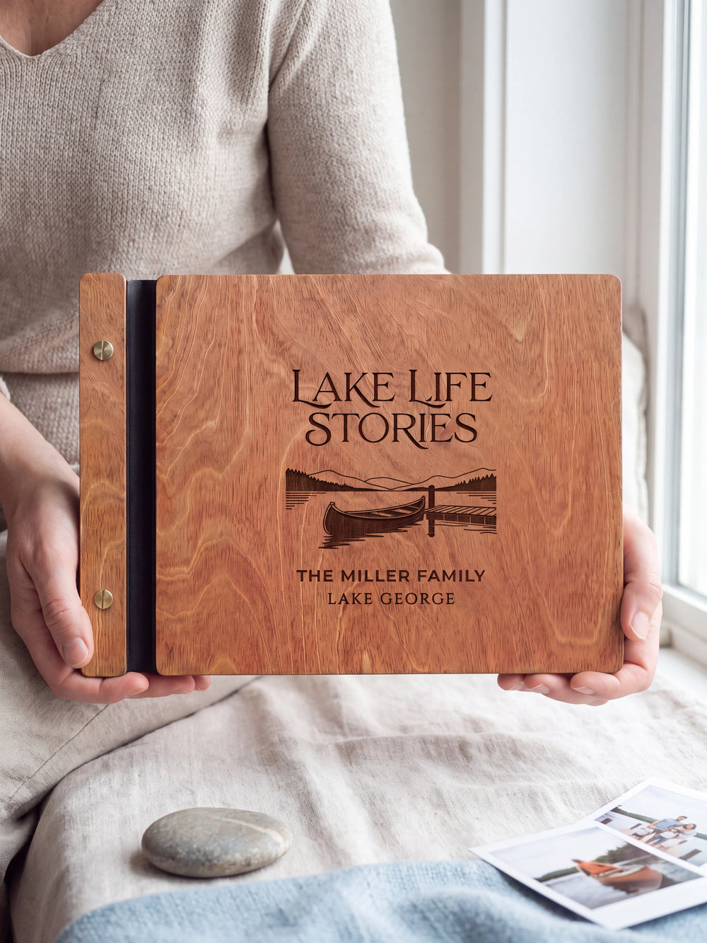 Hands hold the Hazelnut wooden album near a window. The full cover displays a canoe beside a lakeside dock in dark engraving, with personalized lettering and a black binding strip.