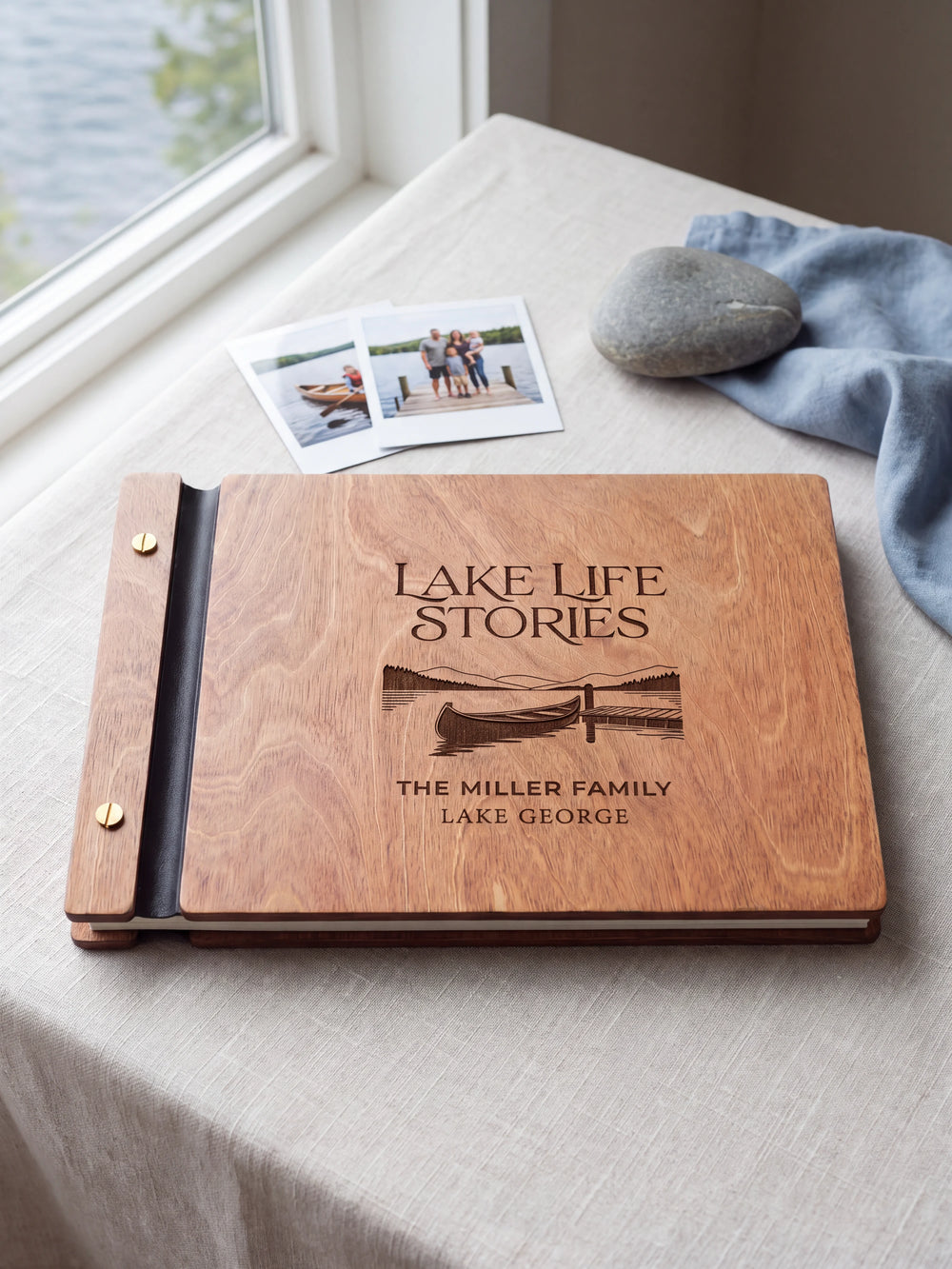 Hazelnut wooden album on a light tabletop with neutral linen. The full cover shows a canoe beside a lakeside dock, personalized wording and the two-screw binding.