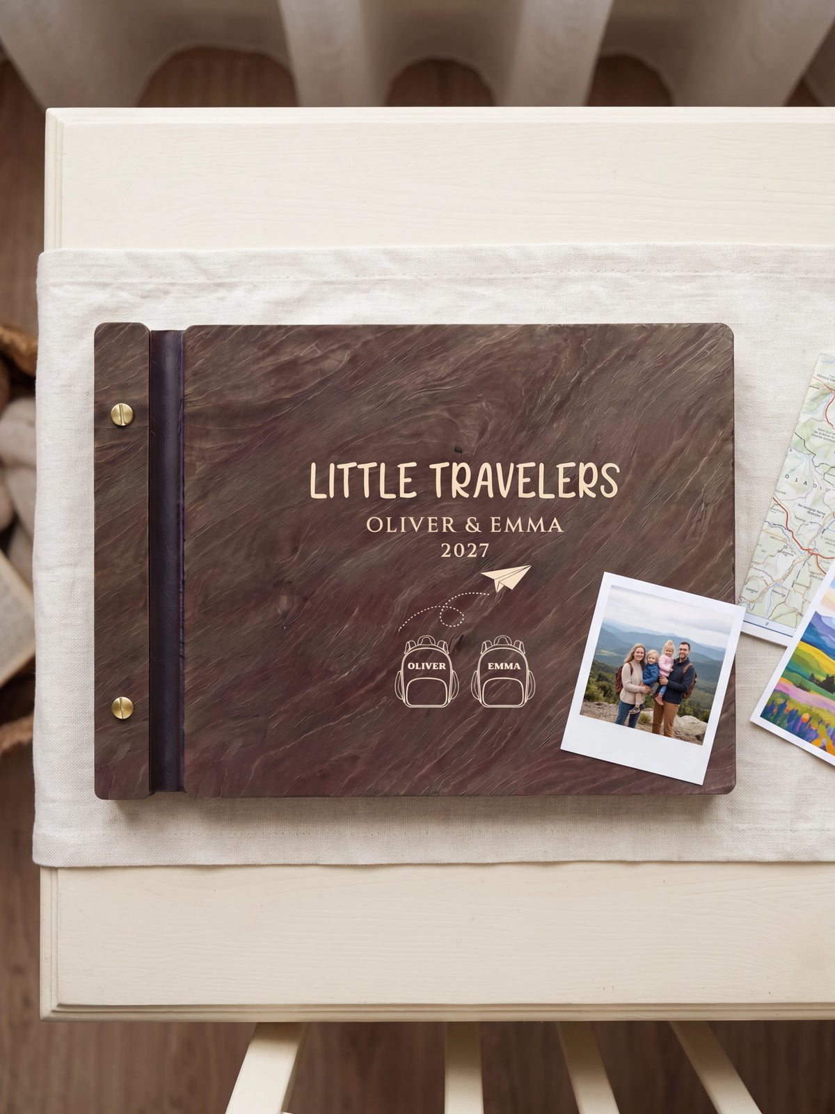 Dark brown Jacobean wooden album on a light surface, with cream engraving of two named backpacks and a paper airplane and personalized cover wording. The full cover and left screw binding are visible.