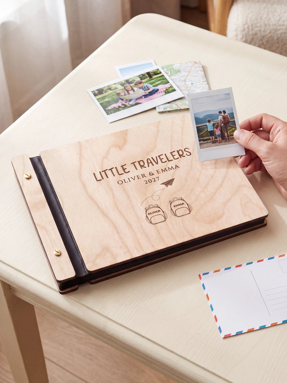 Natural wooden album with dark engraving of two named backpacks and a paper airplane. A hand holds an instant photo beside the closed cover, with loose snapshots nearby.