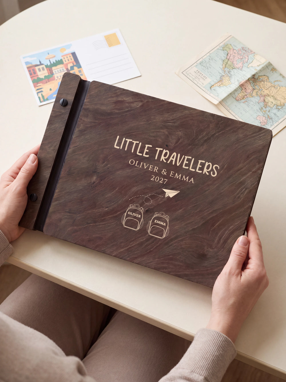 Hands hold a dark brown Jacobean wooden album close to the camera. Cream engraving shows two named backpacks and a paper airplane, personalized wording and the black binding strip.