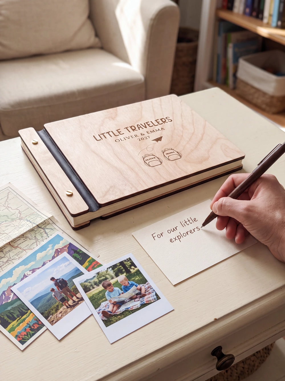 A Natural wooden album showing two named backpacks and a paper airplane rests beside a separate gift card. A hand writes 
