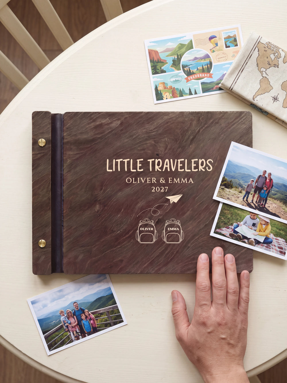 A hand rests beside the closed dark brown Jacobean album on a table. Cream engraving and personalized wording are visible, with travel photos nearby.