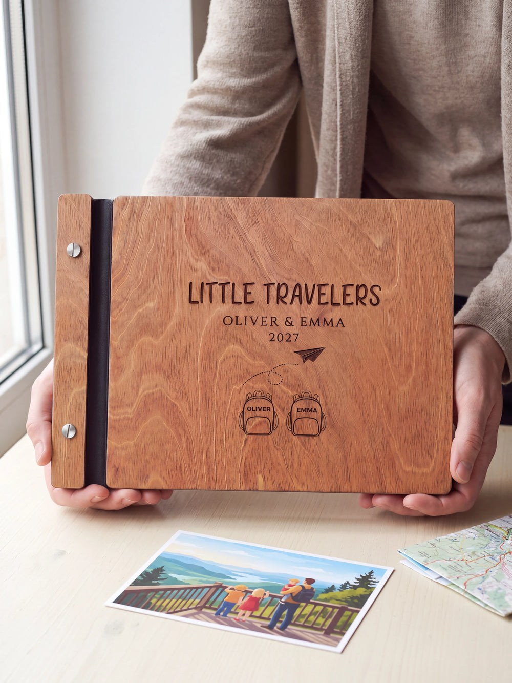 Hands hold the Hazelnut wooden album near a window. The full cover displays two named backpacks and a paper airplane in dark engraving, with personalized lettering and a black binding strip.