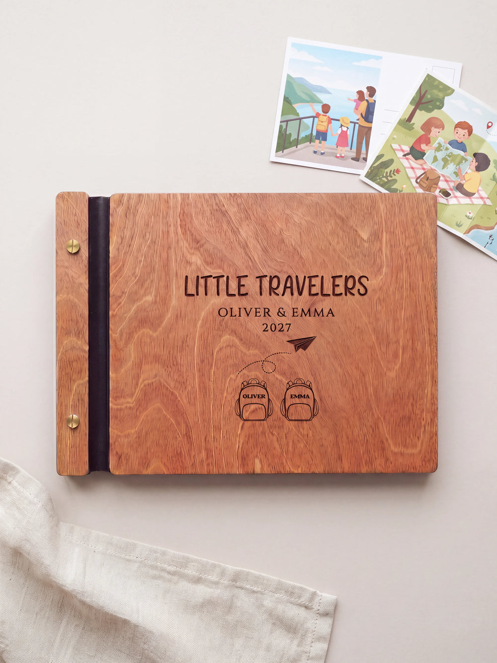 Hazelnut wooden album on a light tabletop with neutral linen. The full cover shows two named backpacks and a paper airplane, personalized wording and the two-screw binding.