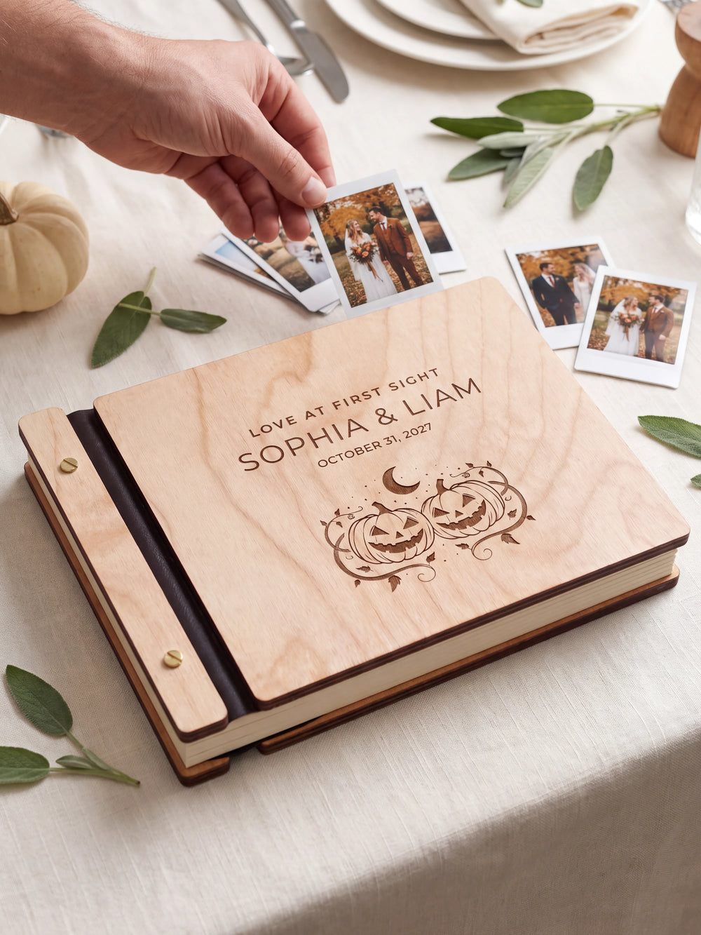 Natural wood Love at First Sight photo guest book on a light table while a hand picks an instant photo to add; the engraved cover reads “LOVE AT FIRST SIGHT”, “SOPHIA & LIAM” and “OCTOBER 31, 2027”