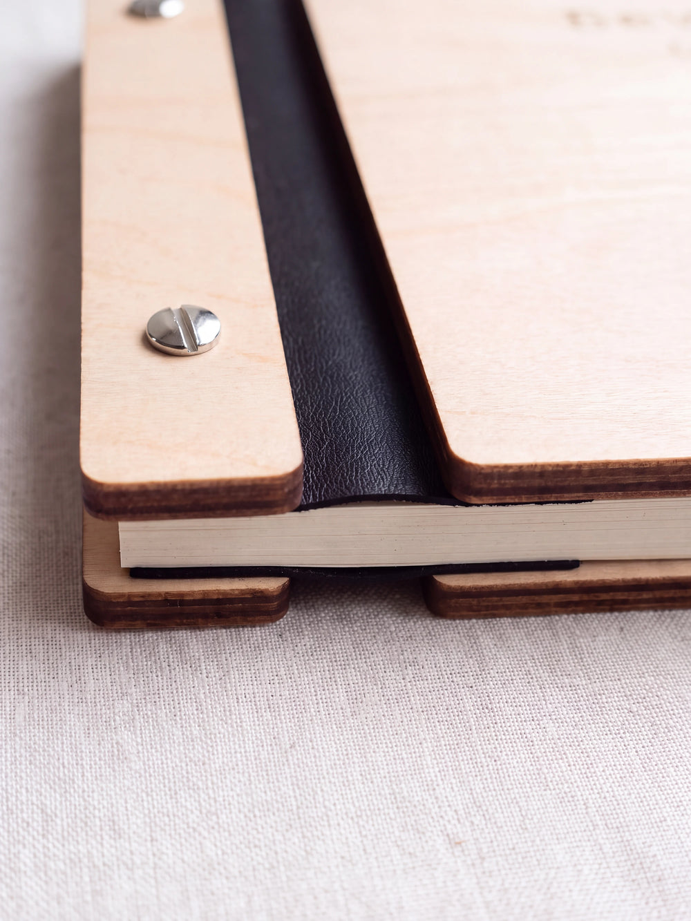 Close-up of a polished silver screw in the Natural wood strip beside the black vegan leather binding, the cream sheet stack visible below