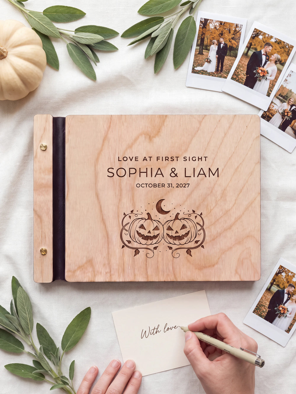 A hand writing a small gift card beside the closed Natural wood Love at First Sight photo guest book; the engraved cover and silver screws are visible