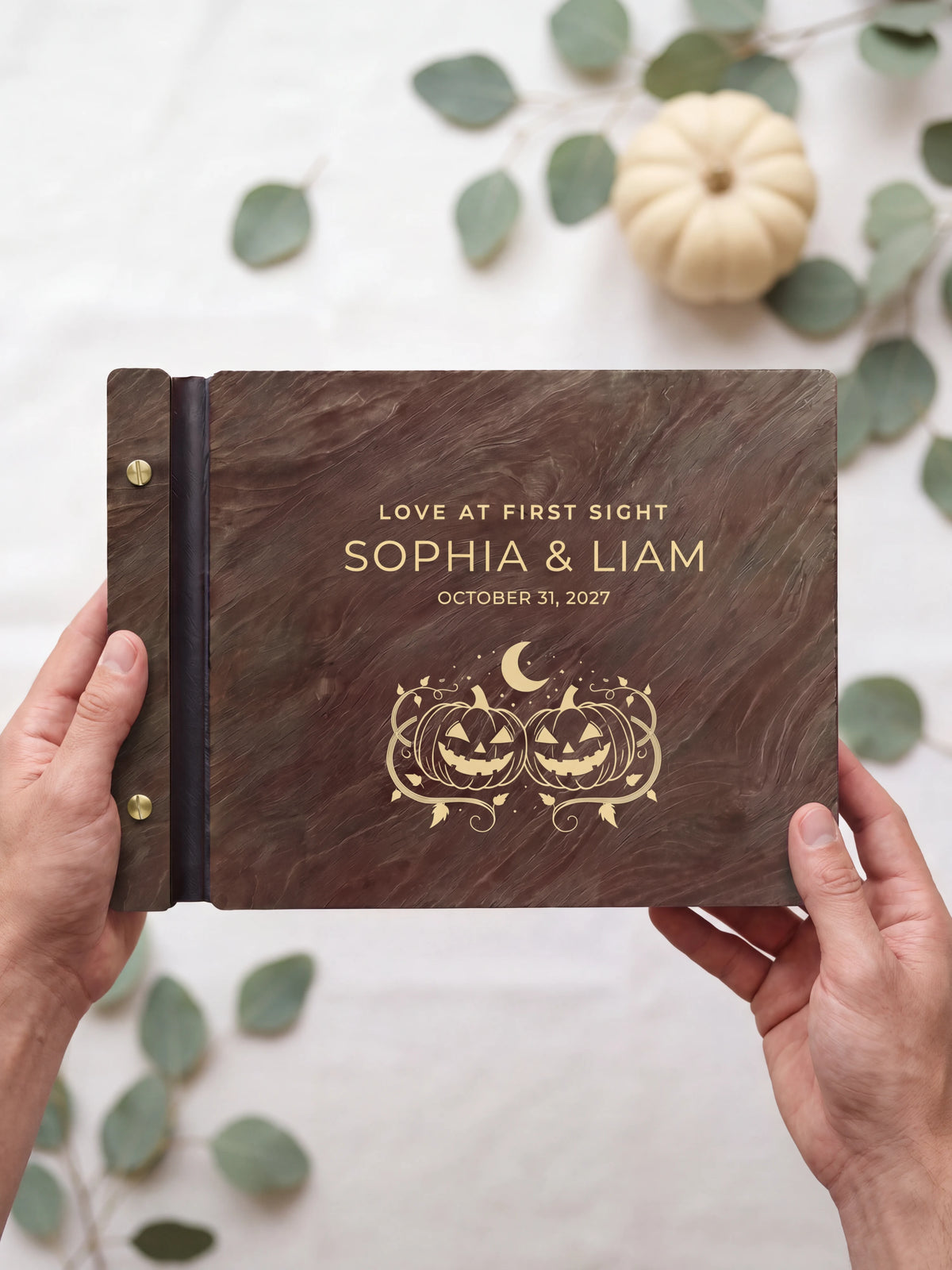 Two hands presenting the closed Jacobean Love at First Sight photo guest book with its gold-tone engraving of two smiling carved pumpkins under a crescent moon, sage leaves and a small pale pumpkin beside them
