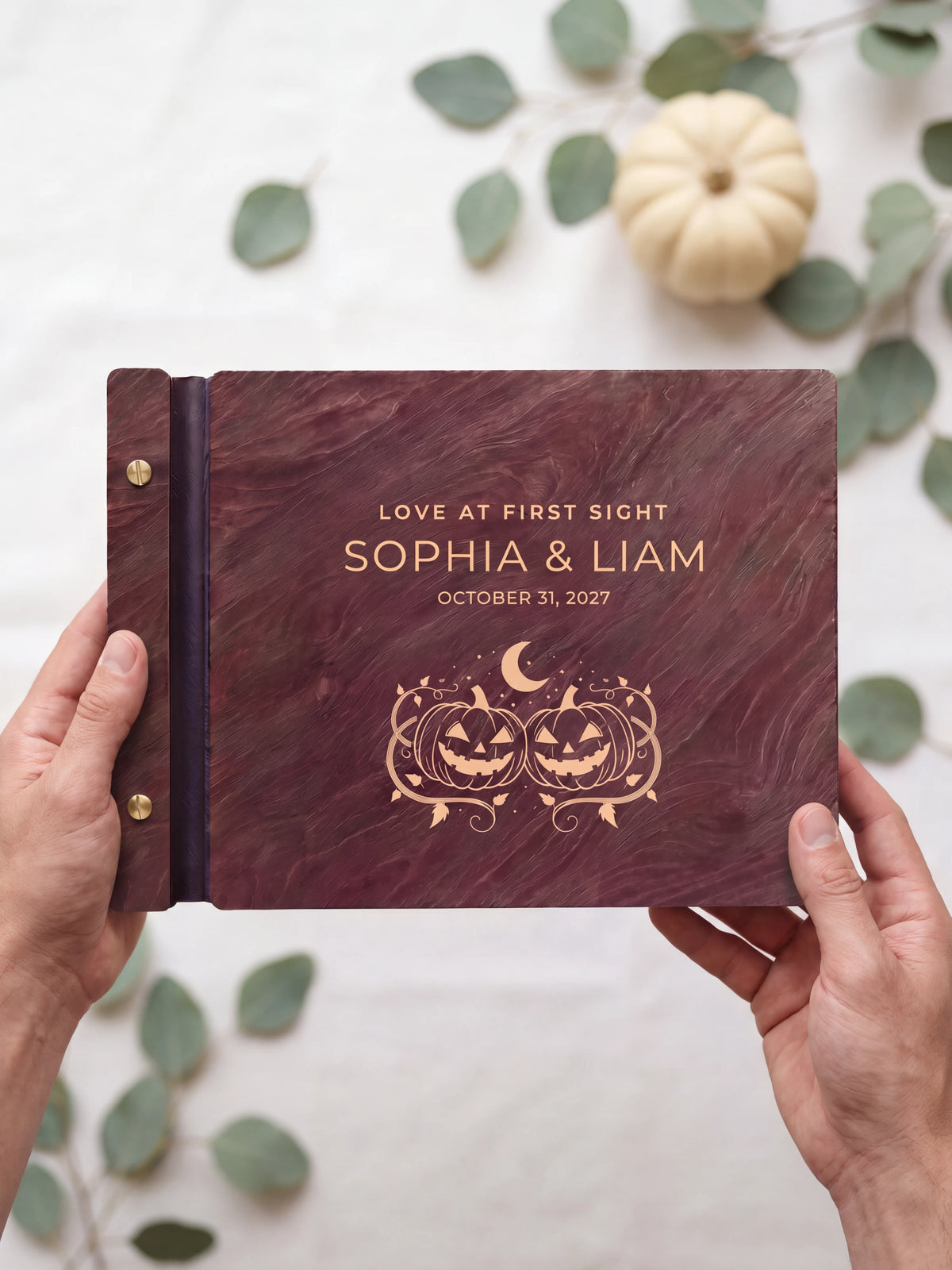 Two hands presenting the closed Jacobean Love at First Sight photo guest book with its cream engraving of two smiling carved pumpkins under a crescent moon, sage leaves and a small pale pumpkin beside them