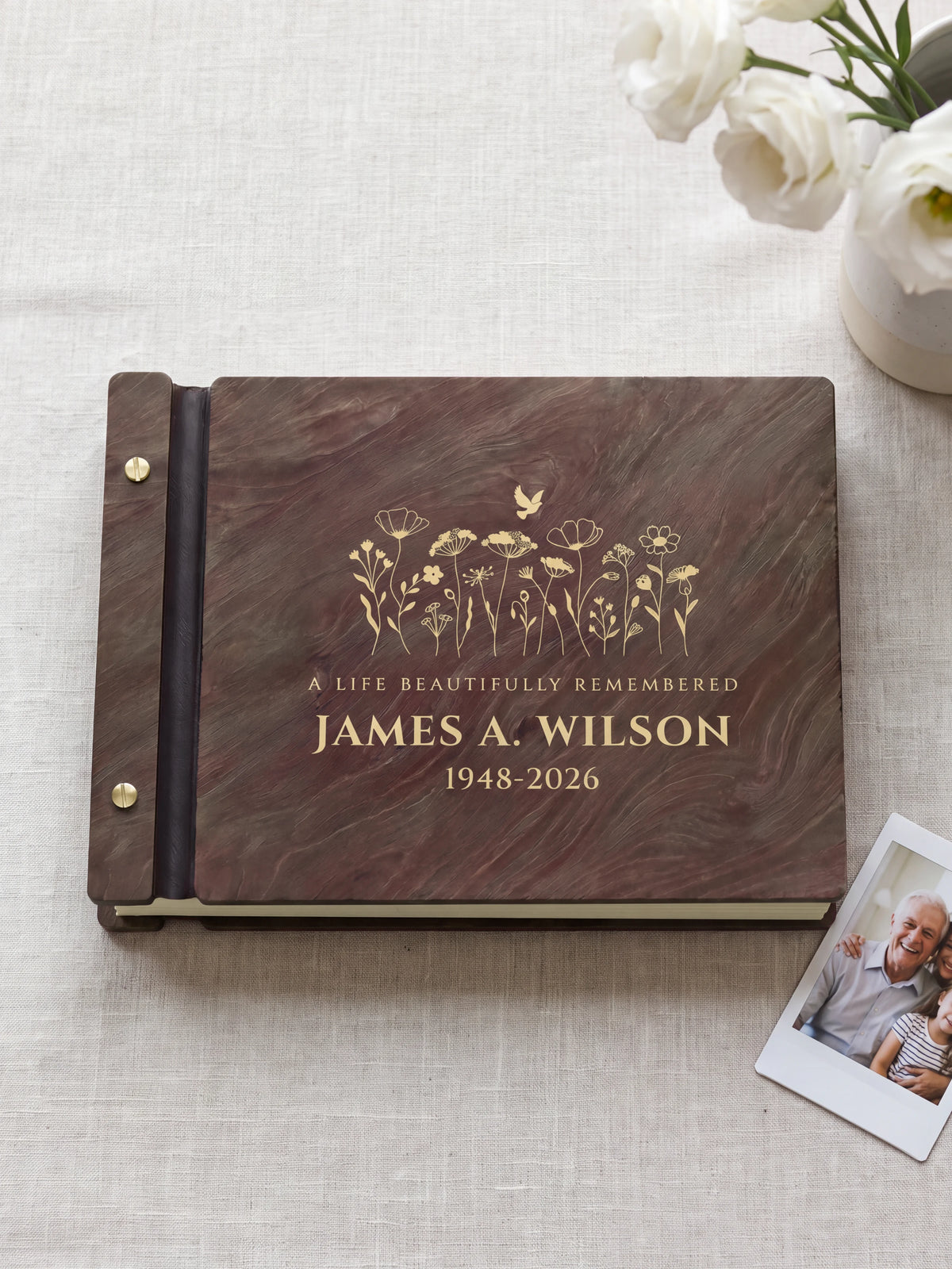 Jacobean wood memorial photo guest book lying flat on gray linen, the cover engraved with “A LIFE BEAUTIFULLY REMEMBERED”, “JAMES A. WILSON” and “1948-2026”; two silver screws along the black vegan leather spine