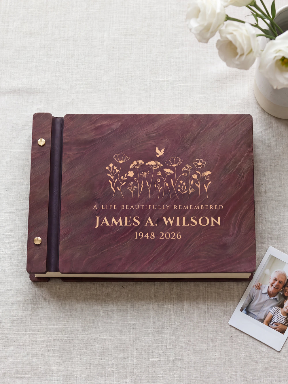 Jacobean wood memorial photo guest book lying flat on gray linen, the cover engraved with “A LIFE BEAUTIFULLY REMEMBERED”, “JAMES A. WILSON” and “1948-2026”; two silver screws along the black vegan leather spine