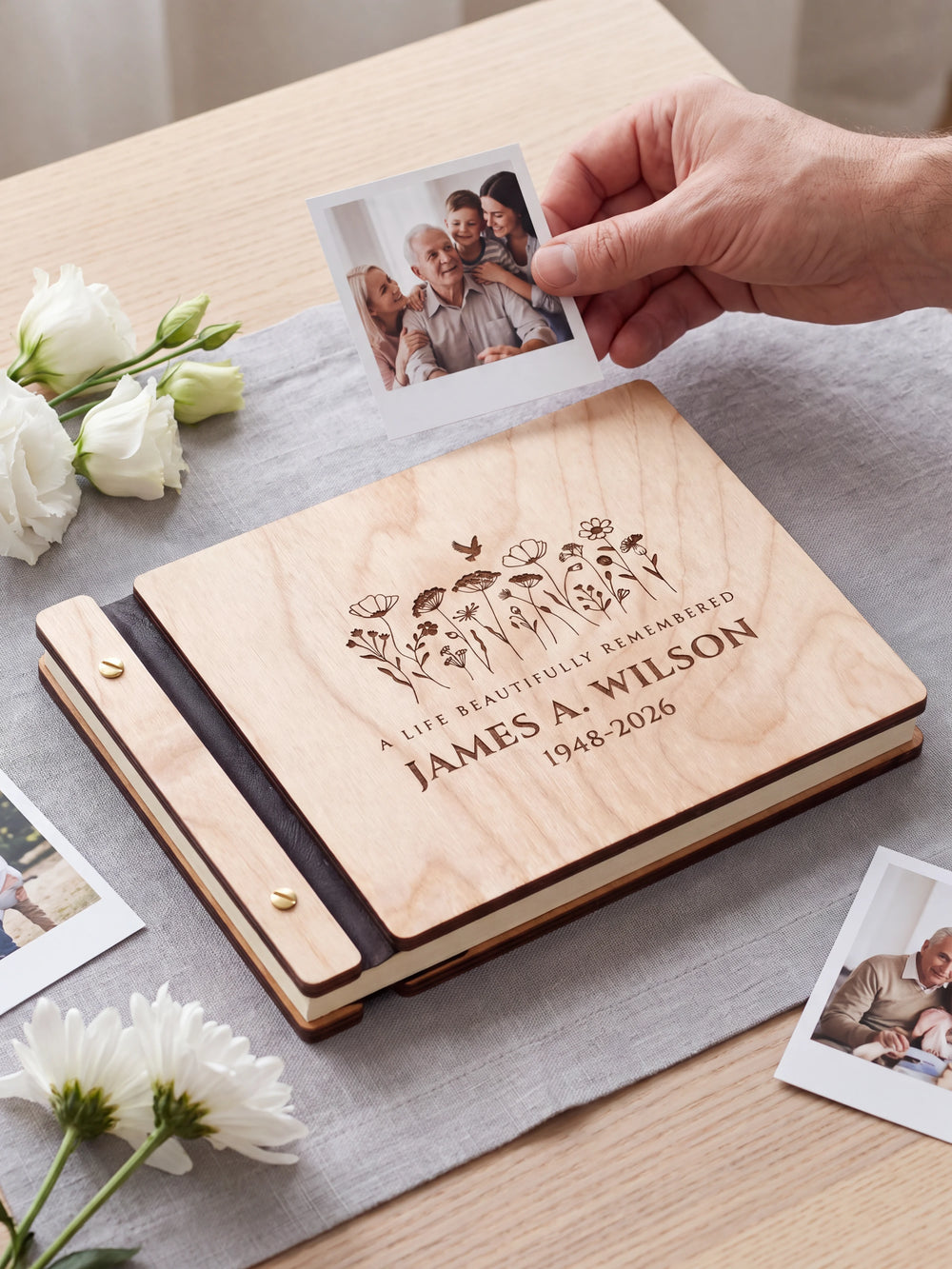 Natural wood memorial photo guest book on a light table while a hand picks an instant photo to add; the engraved cover reads “A LIFE BEAUTIFULLY REMEMBERED”, “JAMES A. WILSON” and “1948-2026”