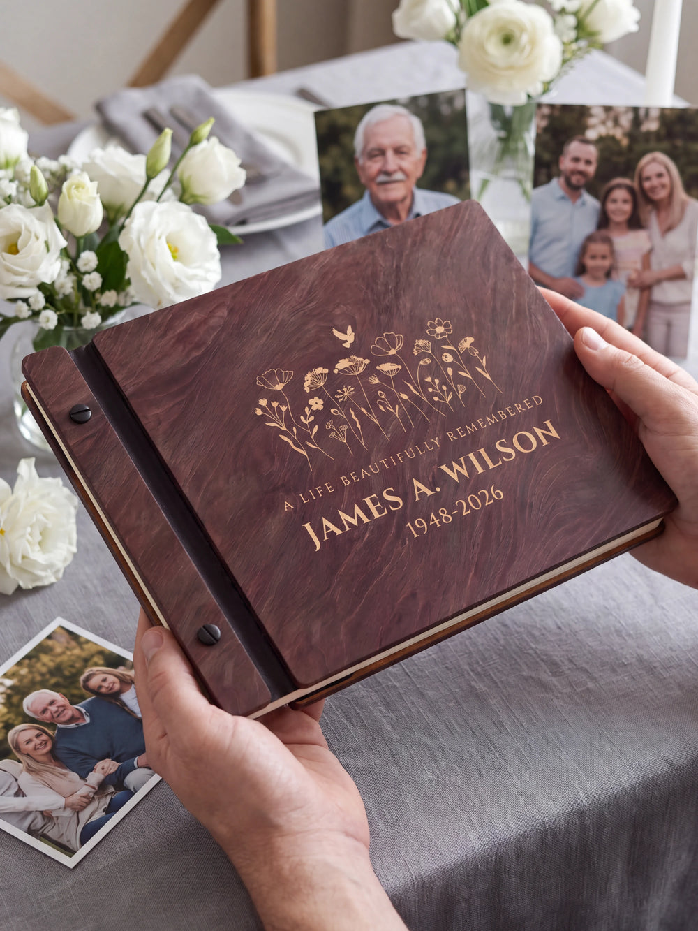 Two hands holding the Jacobean memorial photo guest book up to the camera, the cream engraving of a wildflower meadow with a small dove and “A LIFE BEAUTIFULLY REMEMBERED”, “JAMES A. WILSON” and “1948-2026” filling the cover