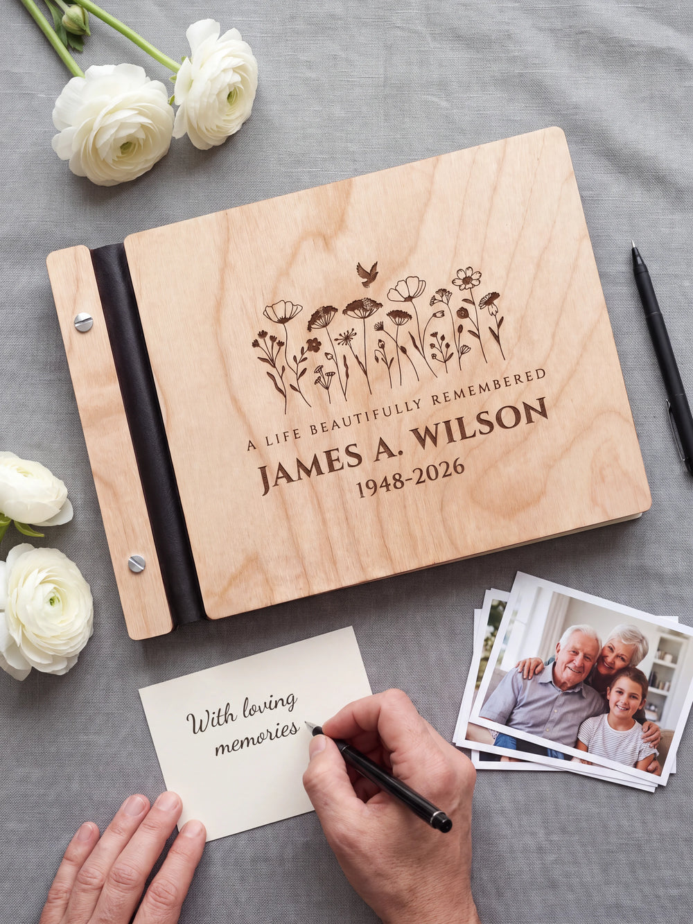 A hand writing a small gift card beside the closed Natural wood memorial photo guest book; the engraved cover and silver screws are visible
