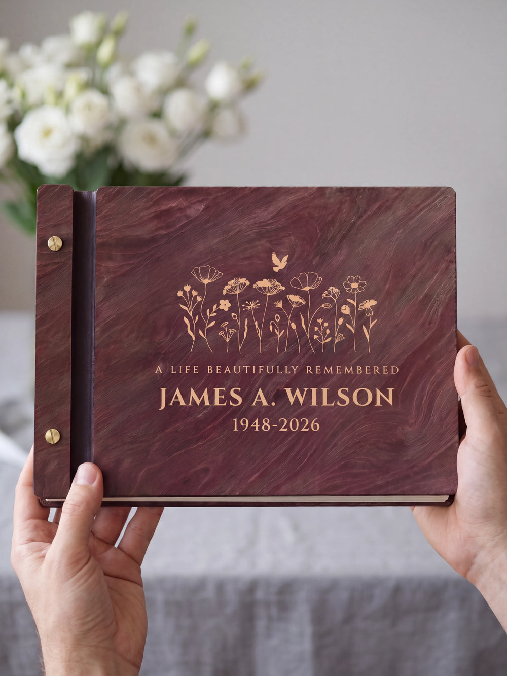 Two hands presenting the closed Jacobean memorial photo guest book with its cream engraving of a wildflower meadow with a small dove, white flowers on the table beside them