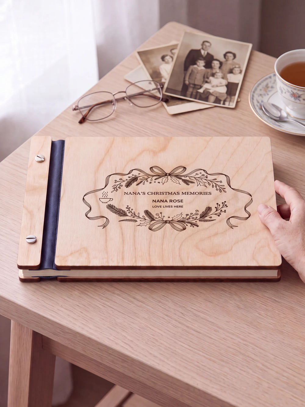 Natural wooden album with brown laser engraving of ribbon oval wreath, foliage and a small steaming cup, shown in a close lifestyle view with navy blue binding and silver screws. The cover reads NANA’S CHRISTMAS MEMORIES.