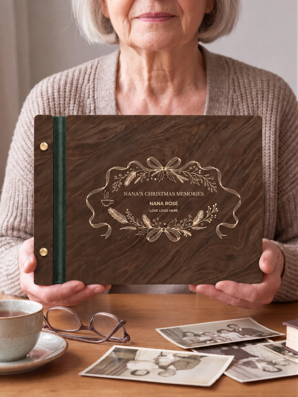 Two hands hold a Jacobean wooden photo album with the NANA’S CHRISTMAS MEMORIES cover facing the camera. Cream engraved artwork, the narrow binding and gold screws are visible.