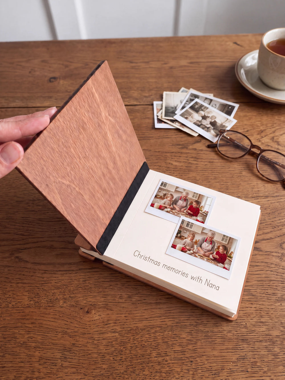 A hand lifts the Hazelnut album’s front cover, revealing a flat black inner fabric strip and a cream page arranged with instant photographs and a handwritten memory.