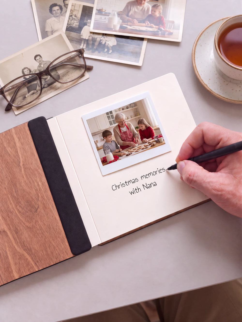 An open Hazelnut album with a photograph on a cream page. A hand writes a memory beneath it; a flat black fabric strip runs along the inner wooden cover.