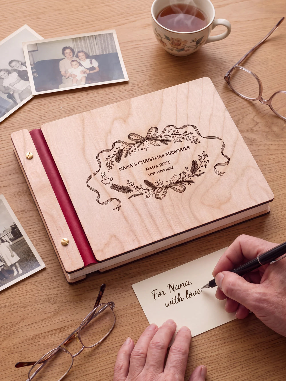 A Natural wooden album with the NANA’S CHRISTMAS MEMORIES engraving, cherry red binding and gold screws. A hand writes on a separate gift card beside the book.