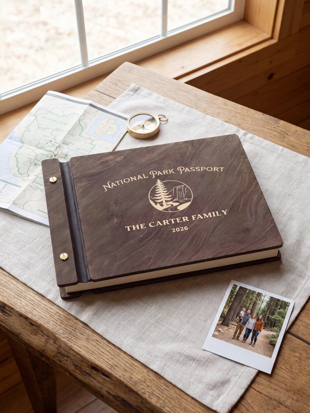 Dark brown Jacobean wooden album on a light surface, with cream engraving of a pine tree and rocky park landscape and personalized cover wording. The full cover and left screw binding are visible.