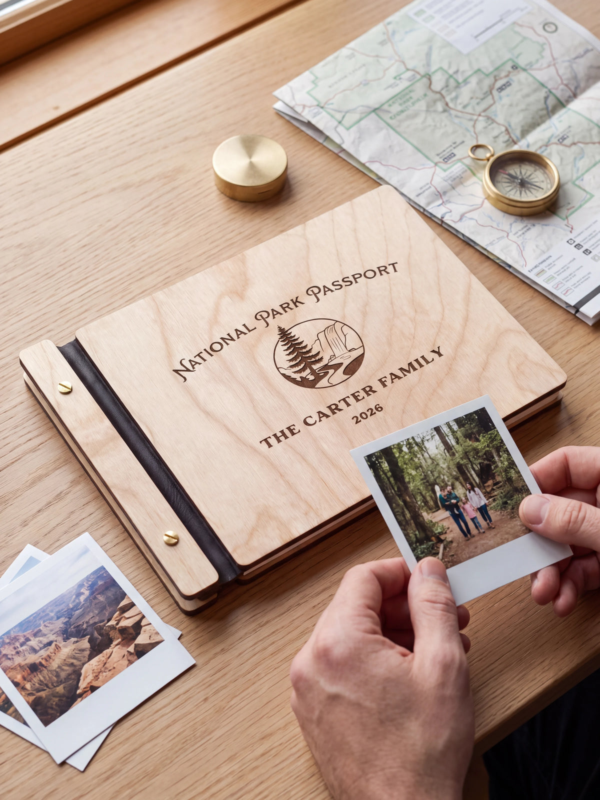 Natural wooden album with dark engraving of a pine tree and rocky park landscape. A hand holds an instant photo beside the closed cover, with loose snapshots nearby.