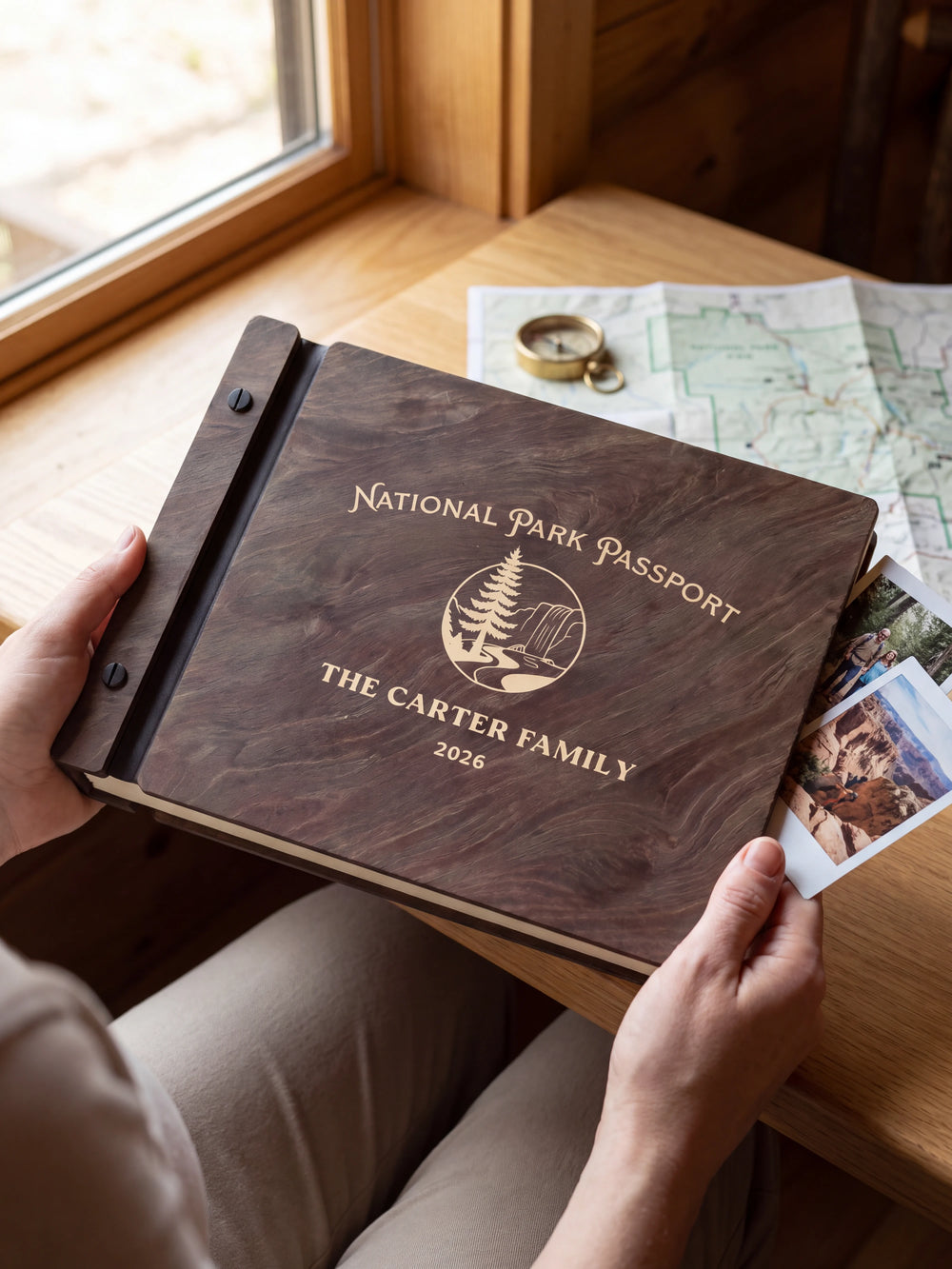 Hands hold a dark brown Jacobean wooden album close to the camera. Cream engraving shows a pine tree and rocky park landscape, personalized wording and the black binding strip.