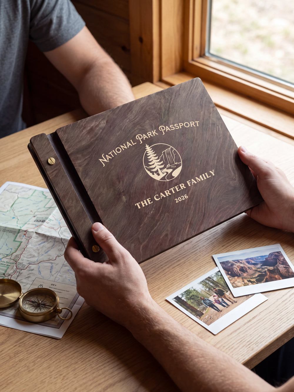 Two hands present a dark brown Jacobean wooden album, with the full cream-engraved pine tree and rocky park landscape design and personalized cover wording facing the camera.