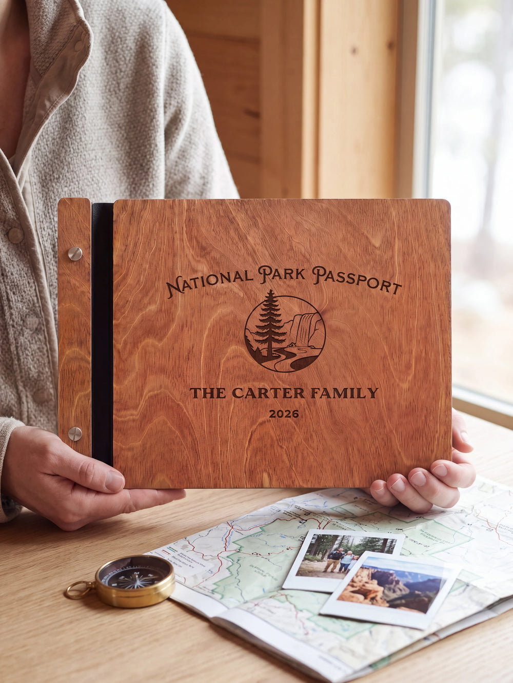 Hands hold the Hazelnut wooden album near a window. The full cover displays a pine tree and rocky park landscape in dark engraving, with personalized lettering and a black binding strip.