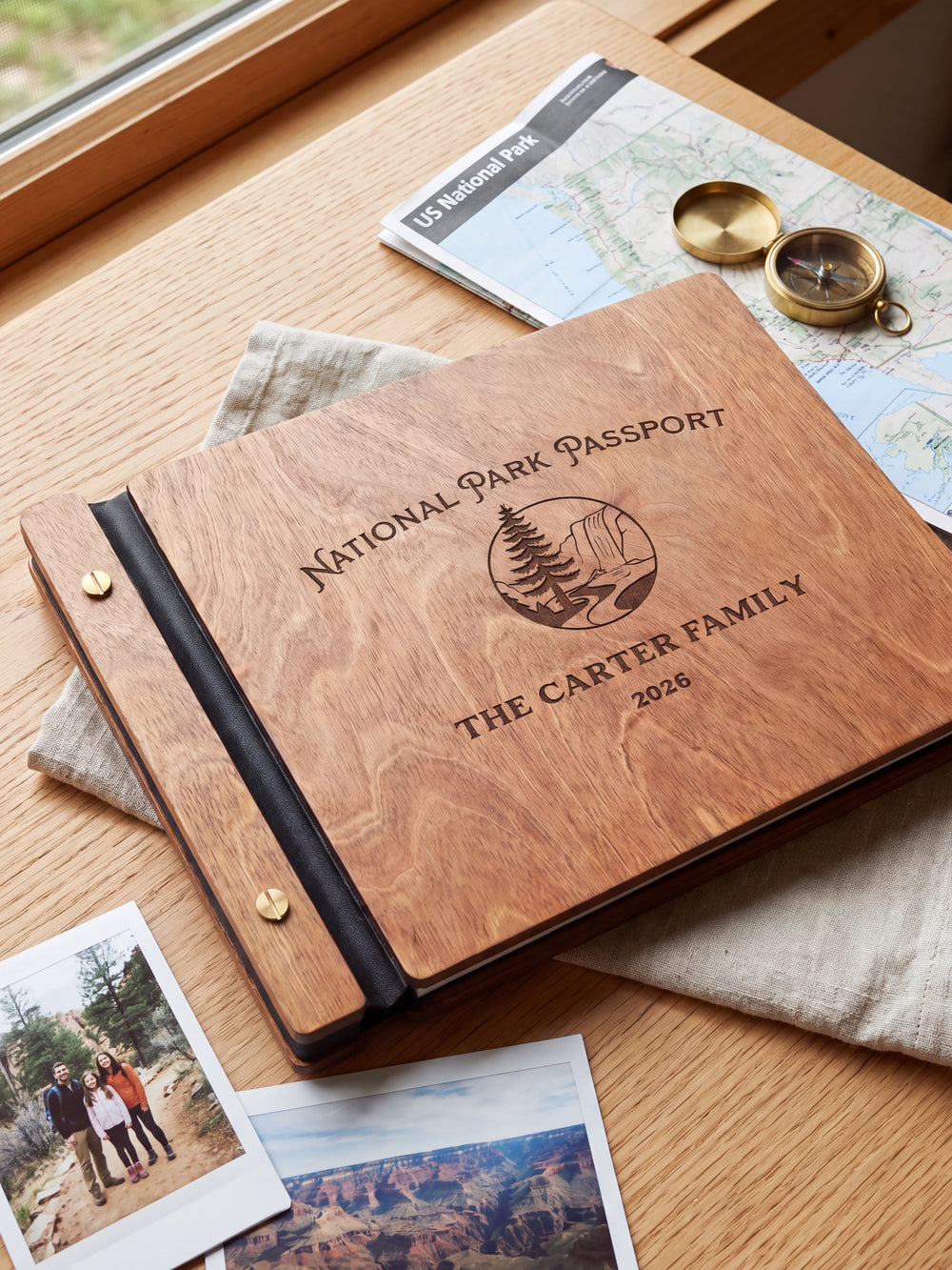 Hazelnut wooden album on a light tabletop with neutral linen. The full cover shows a pine tree and rocky park landscape, personalized wording and the two-screw binding.