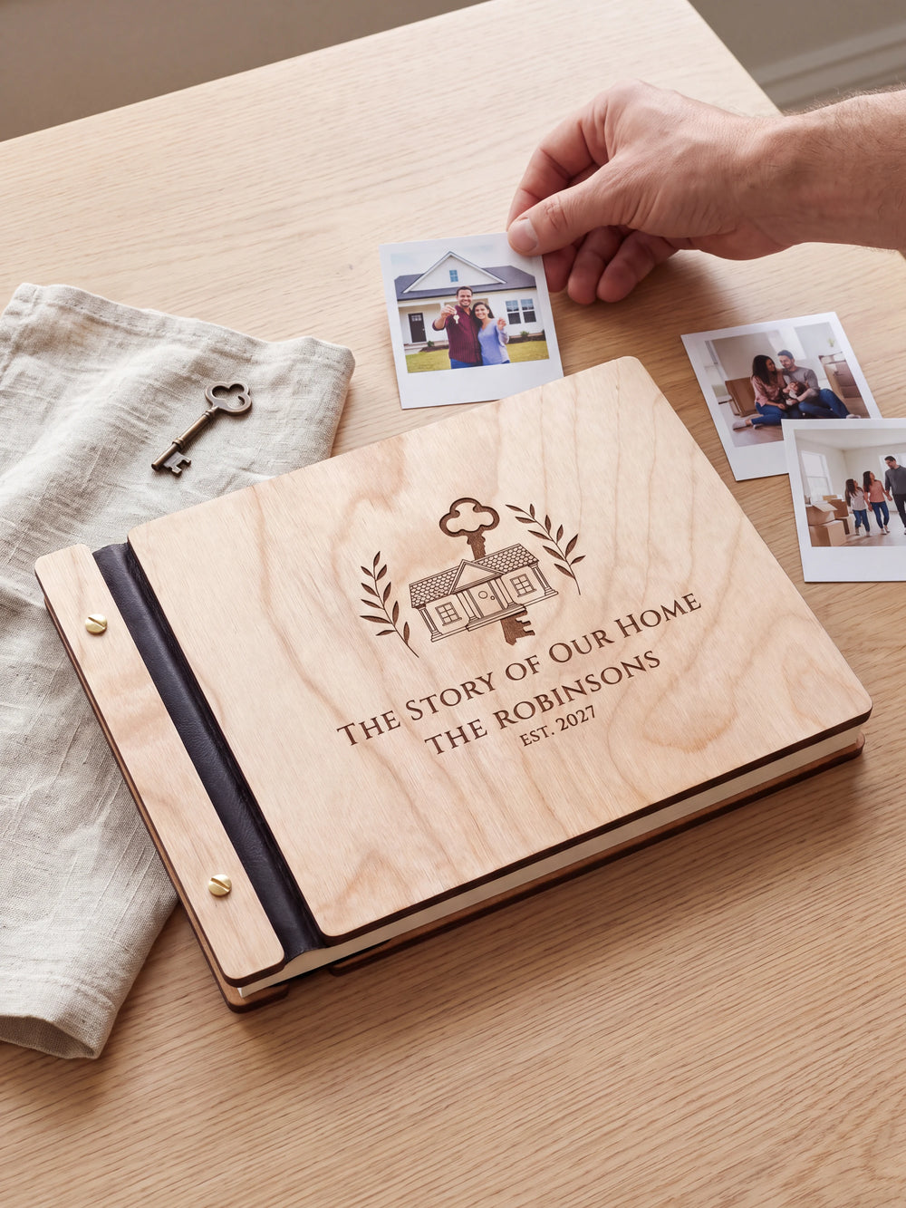 Natural wood The Story of Our Home photo book on a light table while a hand picks an instant photo to add; the engraved cover reads “THE STORY OF OUR HOME”, “THE ROBINSONS” and “EST. 2027”