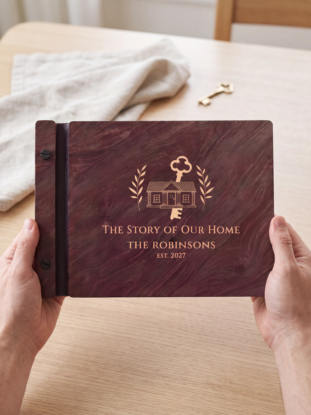 Two hands holding the Jacobean The Story of Our Home photo book up to the camera, the cream engraving of a house with a large old-fashioned key and leafy branches and “THE STORY OF OUR HOME”, “THE ROBINSONS” and “EST. 2027” filling the cover