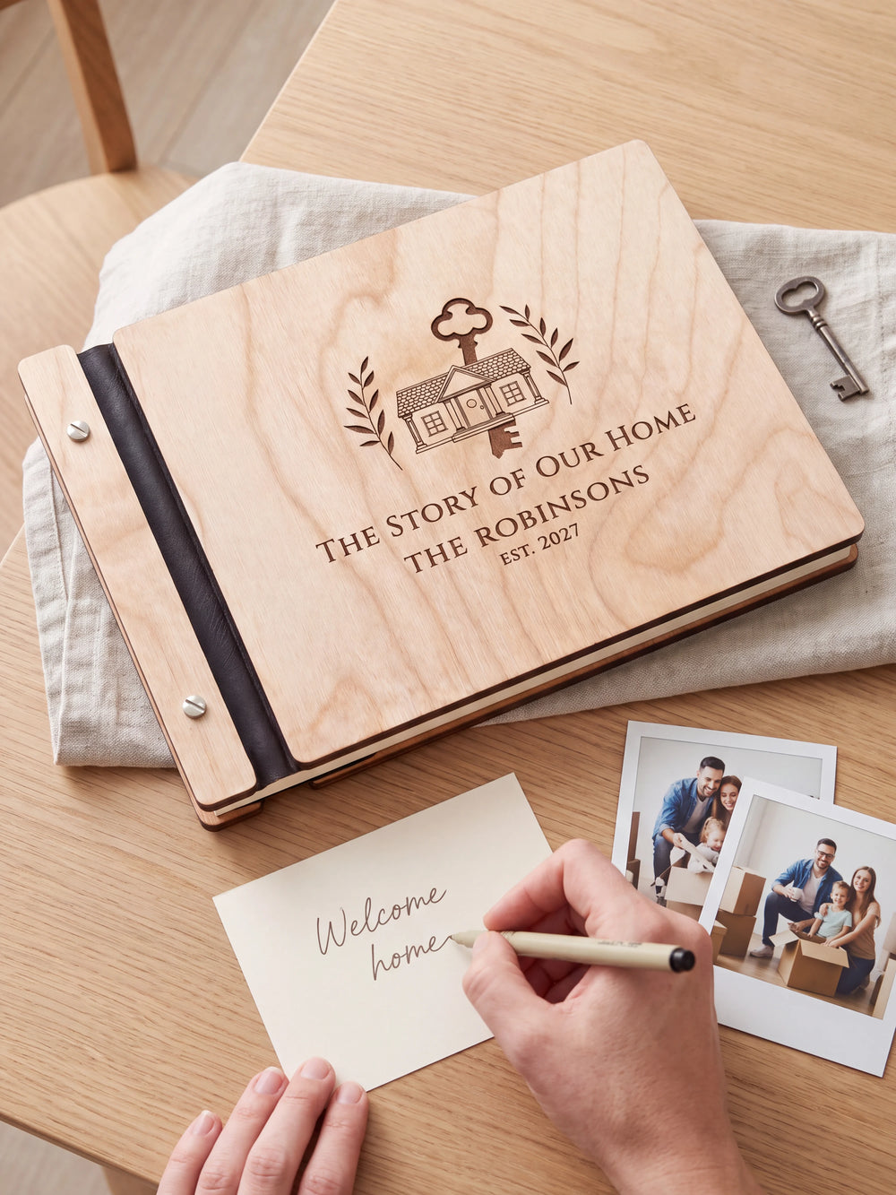 A hand writing a small gift card beside the closed Natural wood The Story of Our Home photo book; the engraved cover and silver screws are visible