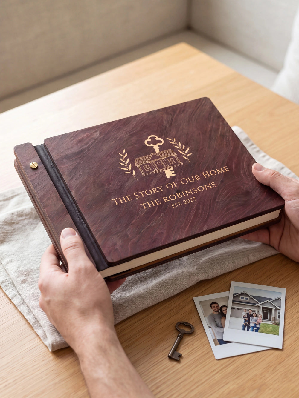 Two hands presenting the closed Jacobean The Story of Our Home photo book with its cream engraving of a house with a large old-fashioned key and leafy branches, a house key on the table beside them
