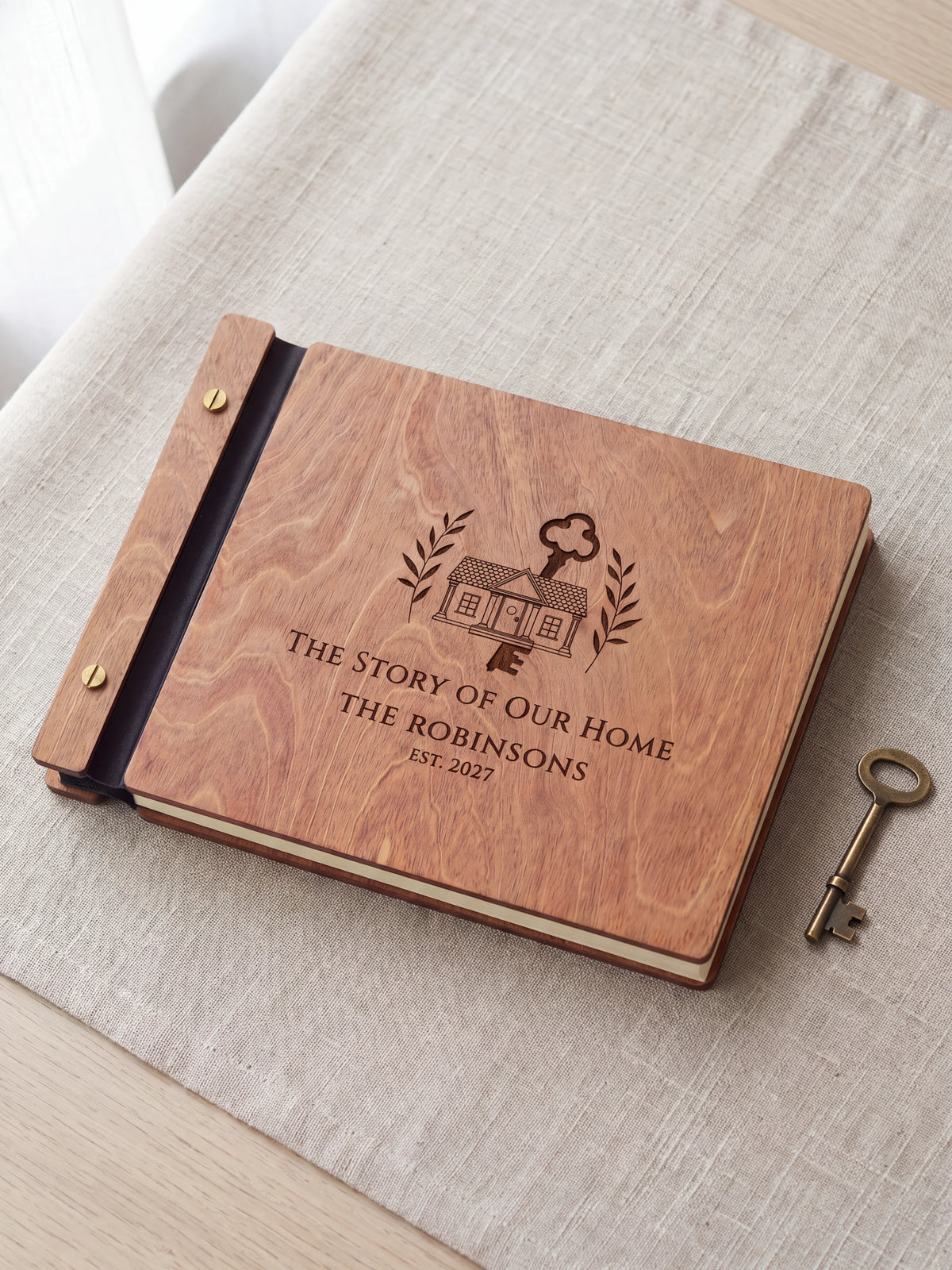 Hazelnut wood The Story of Our Home photo book lying flat on a table, the cover engraved with “THE STORY OF OUR HOME”, “THE ROBINSONS” and “EST. 2027”; two gold-tone screws along the black vegan leather spine