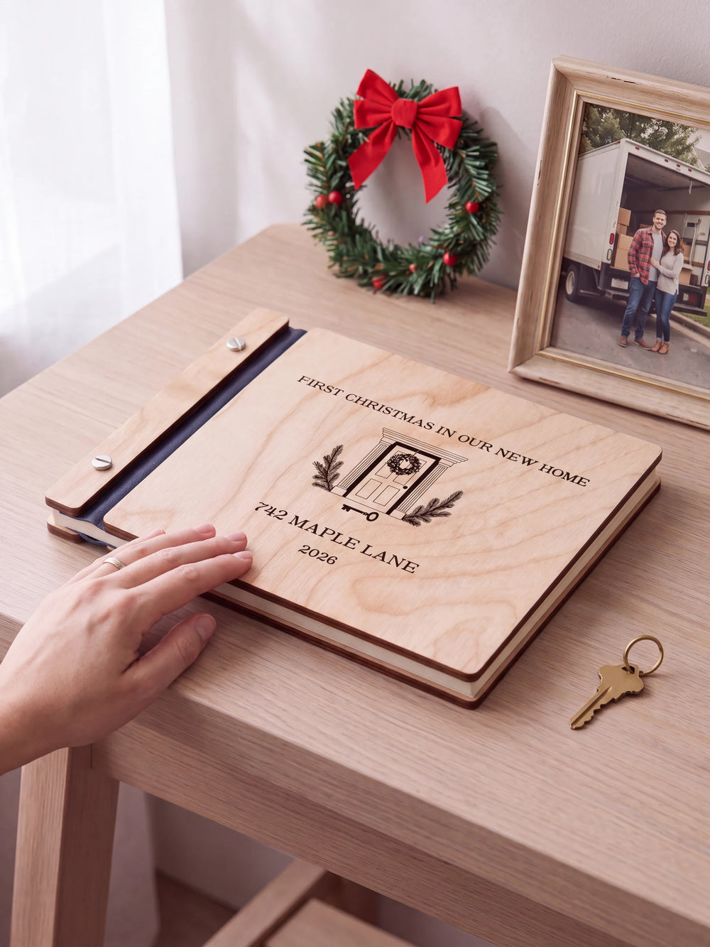 Natural wooden album with brown laser engraving of wreath-adorned front door and house key, shown in a close lifestyle view with navy blue binding and silver screws. The cover reads FIRST CHRISTMAS IN OUR NEW HOME.