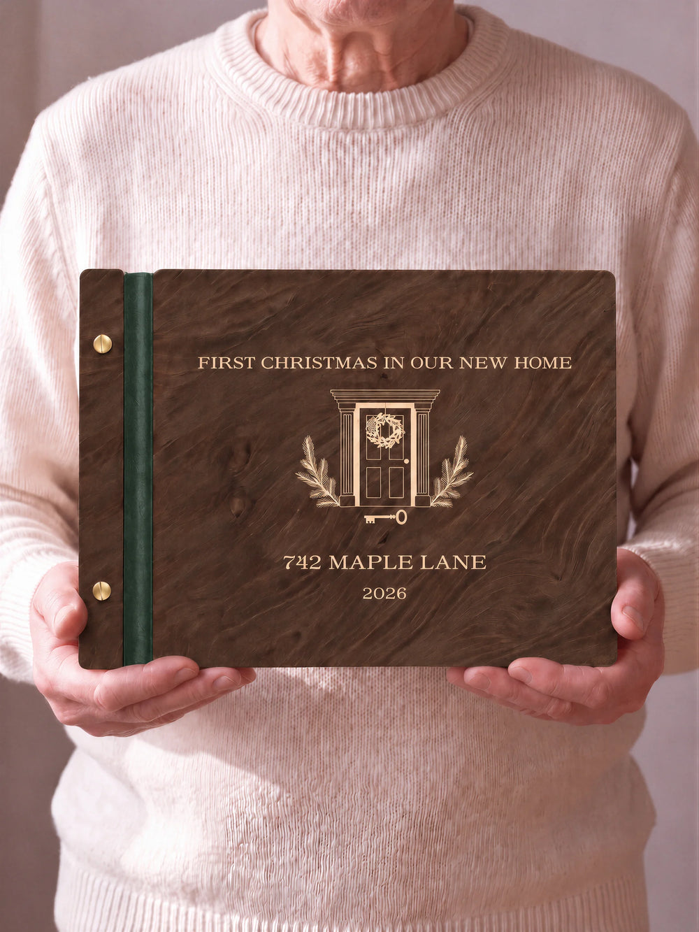 Two hands hold a Jacobean wooden photo album with the FIRST CHRISTMAS IN OUR NEW HOME cover facing the camera. Cream engraved artwork, the narrow binding and gold screws are visible.