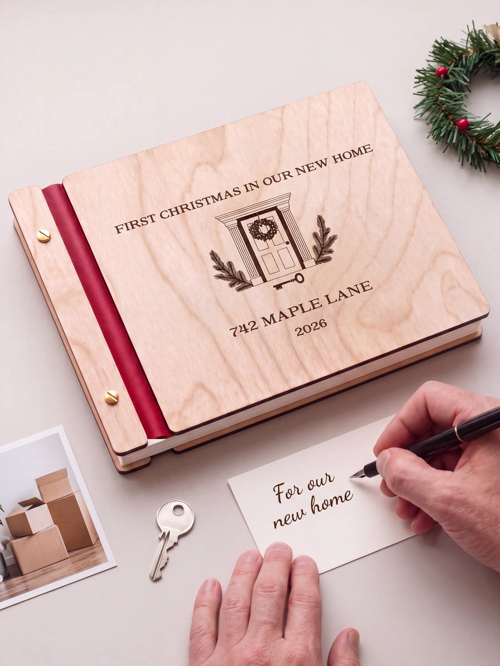 A Natural wooden album with the FIRST CHRISTMAS IN OUR NEW HOME engraving, cherry red binding and gold screws. A hand writes on a separate gift card beside the book.