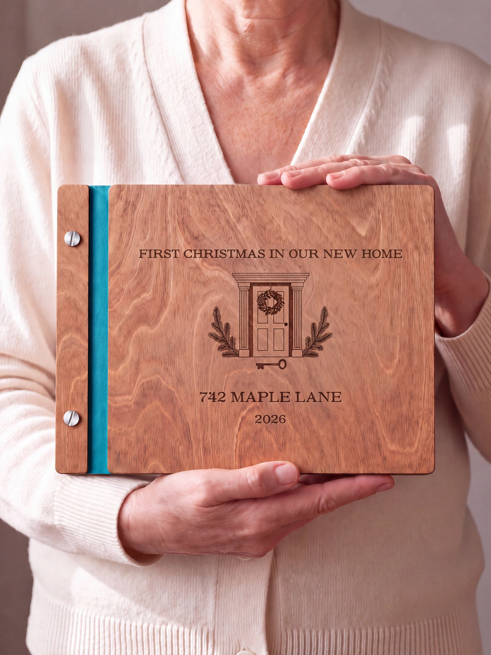 Two hands hold a Hazelnut wooden photo album. The FIRST CHRISTMAS IN OUR NEW HOME cover design has brown engraving, turquoise binding and silver screws.