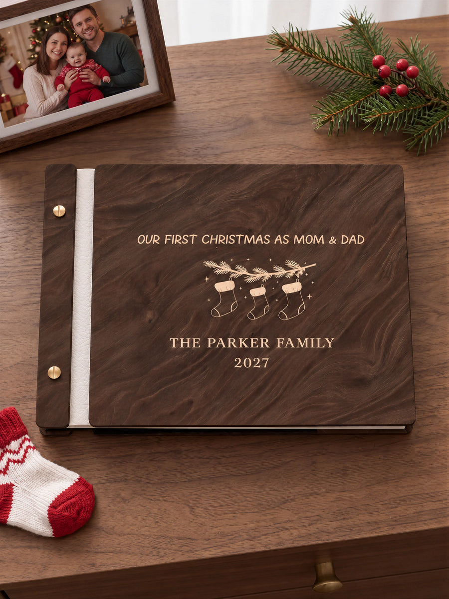 Jacobean wooden album with cream laser engraving of three hanging Christmas stockings and a fir branch. The full personalized cover rests on a styled surface, with ivory binding and gold screws.