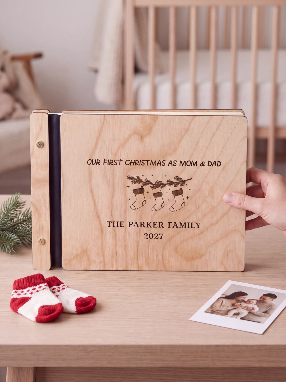 Natural wooden album with brown laser engraving of three hanging Christmas stockings and a fir branch, shown in a close lifestyle view with navy blue binding and silver screws. The cover reads Our First Christmas as Mom & Dad.