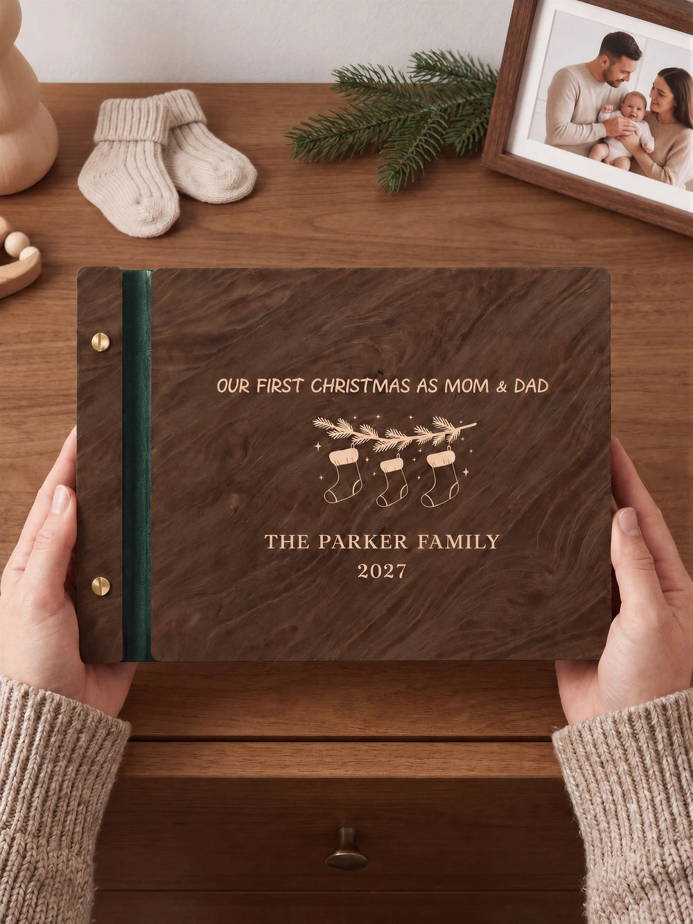 Two hands hold a Jacobean wooden photo album with the Our First Christmas as Mom & Dad cover facing the camera. Cream engraved artwork, the narrow binding and gold screws are visible.