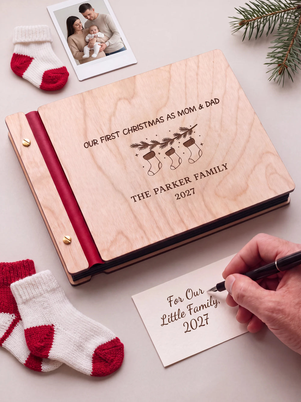 A Natural wooden album with the Our First Christmas as Mom & Dad engraving, cherry red binding and gold screws. A hand writes on a separate gift card beside the book.