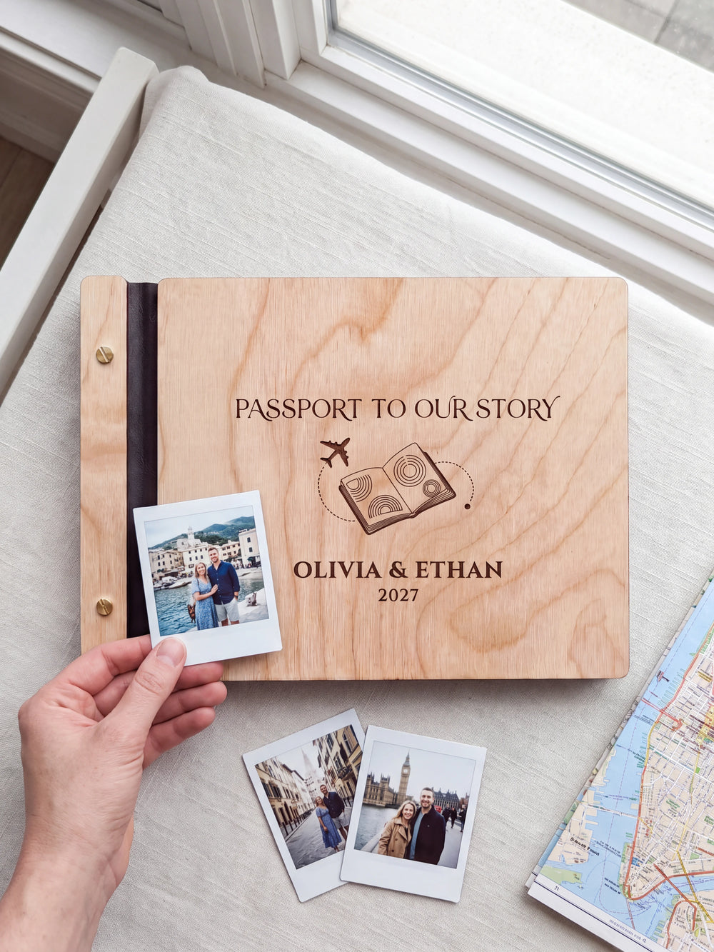 Natural wooden album with dark engraving of a passport and airplane. A hand holds an instant photo beside the closed cover, with loose snapshots nearby.