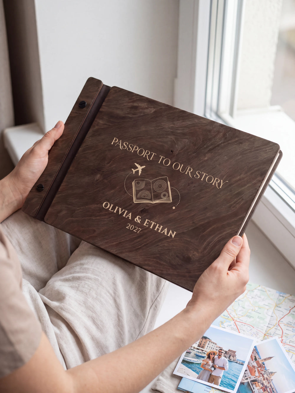 Hands hold a dark brown Jacobean wooden album close to the camera. Cream engraving shows a passport and airplane, personalized wording and the black binding strip.